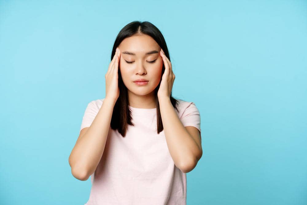 Perempuan muda memegang kedua sisi kepala dengan mata tertutup, tampak kesakitan akibat sakit kepala di latar belakang biru.