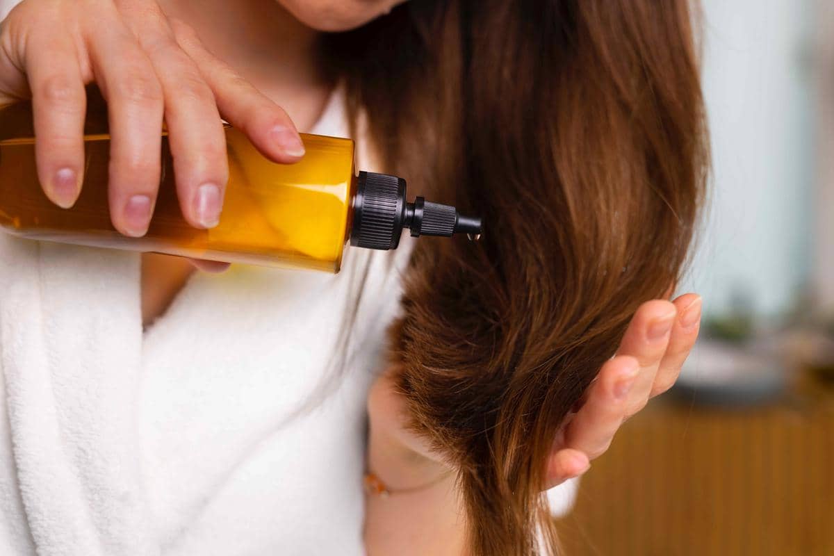 Seseorang mengenakan jubah mandi putih sedang menuangkan minyak perawatan rambut dari botol kuning ke ujung rambut panjangnya.