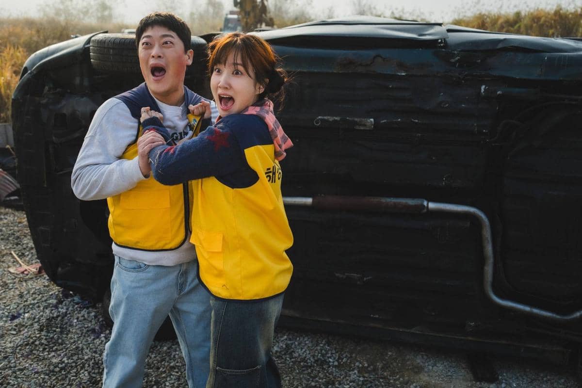 Dua orang mengenakan rompi kuning tampak terkejut di depan mobil terbalik di lokasi luar ruangan pada adegan drama The WONDERfools.