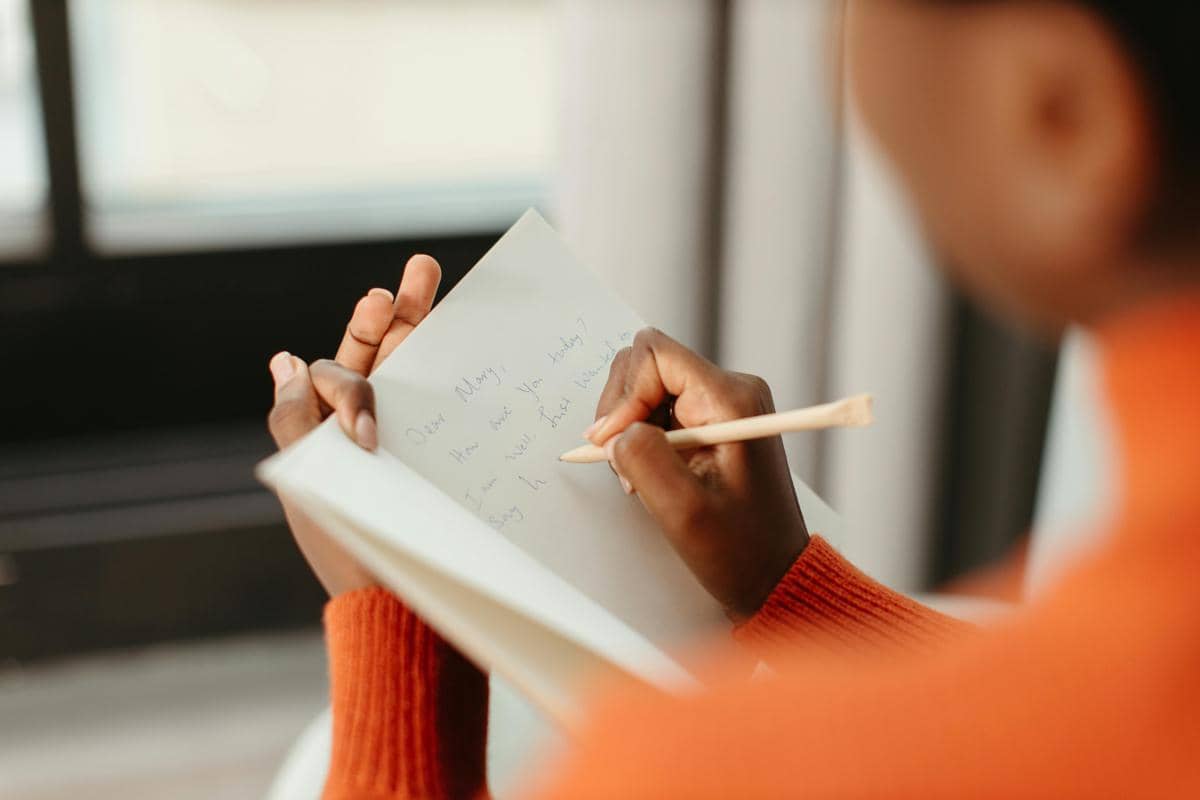 Seseorang mengenakan sweter oranye sedang menulis di buku catatan dengan pensil di dekat jendela yang terang.