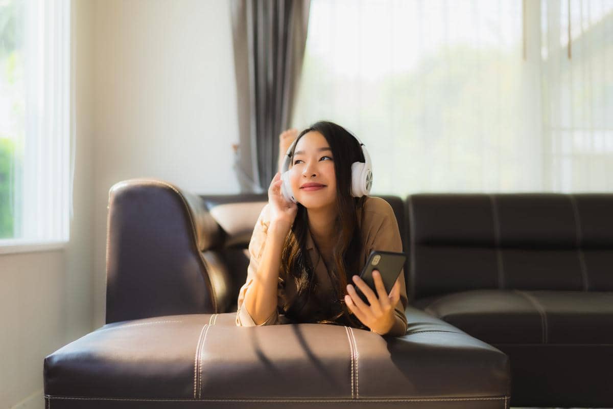Seorang perempuan berbaring santai di sofa sambil mendengarkan musik dengan headphone putih dan memegang ponsel di tangannya.