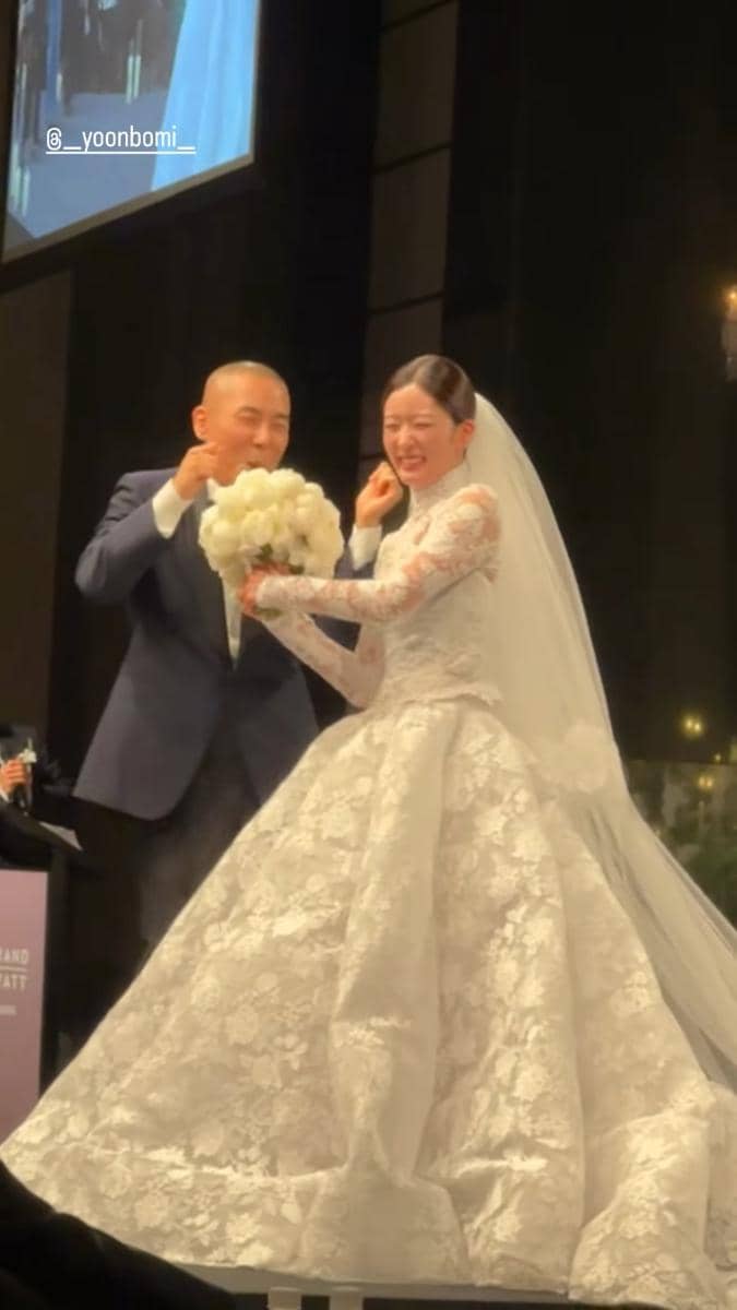 Sepasang pengantin tersenyum bahagia di pelaminan, sang mempelai wanita mengenakan gaun putih panjang dengan detail renda dan membawa buket bunga.