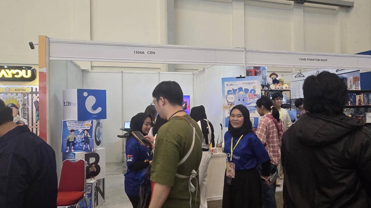 Pengunjung berinteraksi di stan CBN pada acara Comifuro 22 dengan staf mengenakan kaus biru dan latar booth bertema internet fiber.