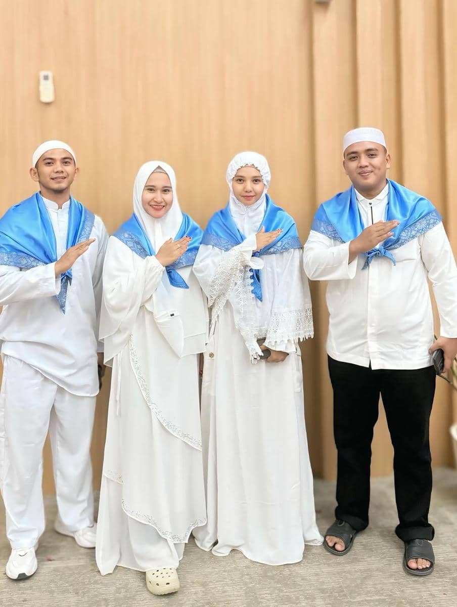 Empat orang mengenakan busana muslim putih dengan selendang biru muda berpose bersama di depan dinding kayu.