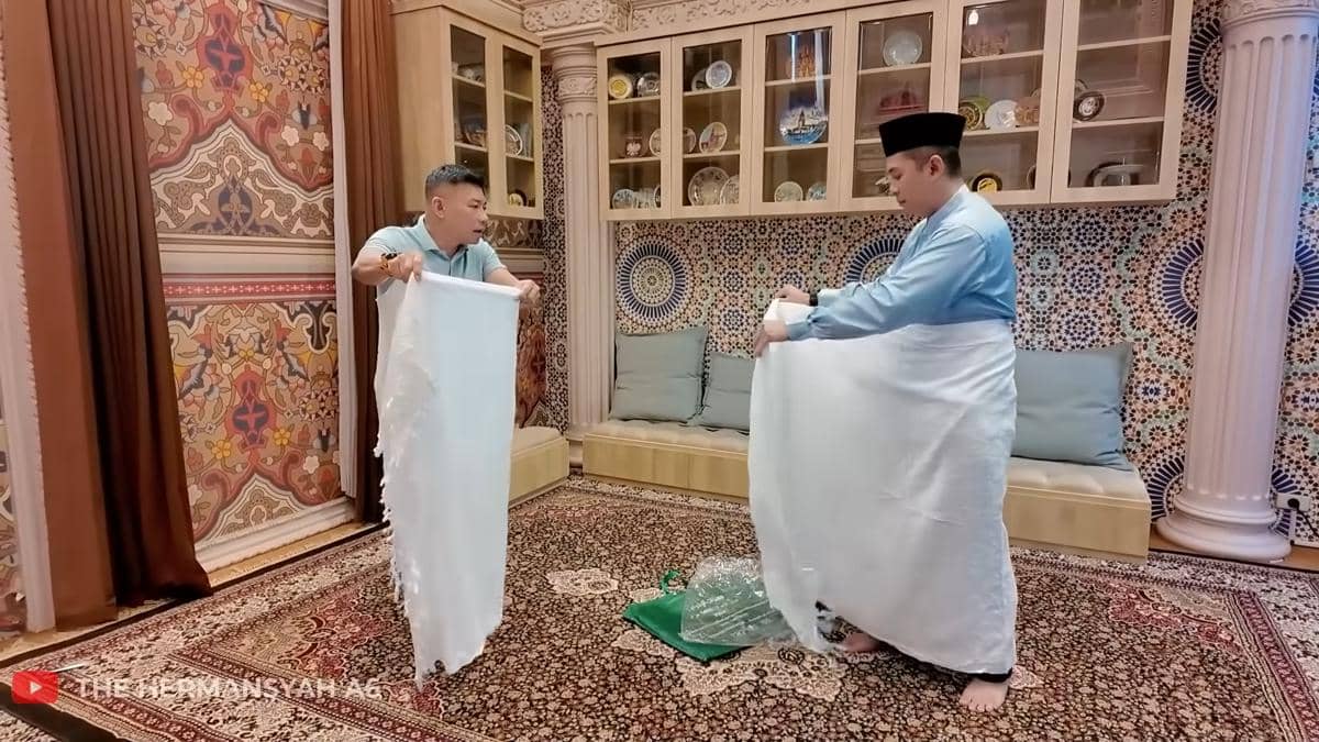 Dua pria di dalam ruangan berpola Timur Tengah sedang memegang kain putih panjang, salah satunya mengenakan baju biru dan peci hitam.