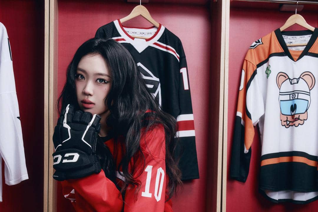 Seorang perempuan berpose mengenakan jersey hoki merah bernomor 10 dan sarung tangan hitam di ruang ganti dengan latar jersey tergantung.