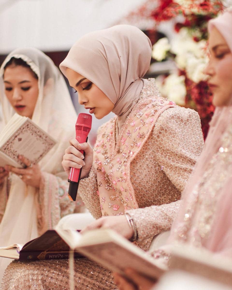 Seorang perempuan berhijab krem memegang mikrofon merah muda dan membaca Al-Qur'an bersama dua perempuan lain dalam acara pengajian.