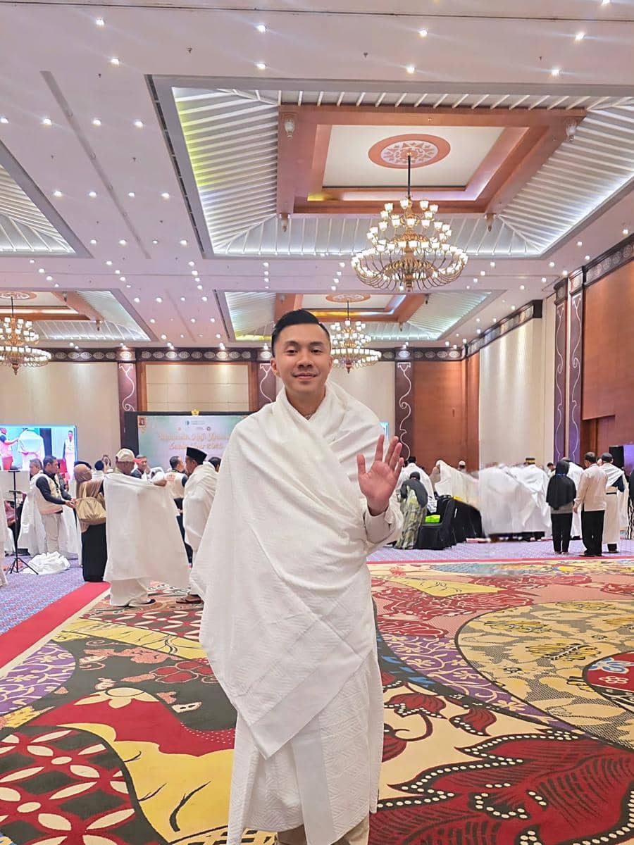 Seorang pria mengenakan kain ihram berwarna putih berdiri di dalam aula besar berhias lampu gantung dan karpet bermotif warna-warni.