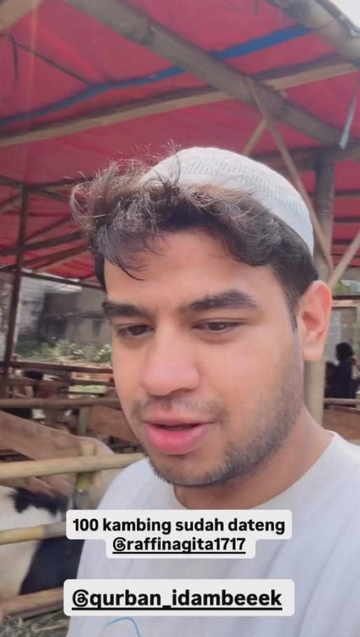 Seorang pria mengenakan peci putih berada di kandang kambing dengan latar belakang atap tenda merah dan beberapa kambing di sekitarnya.