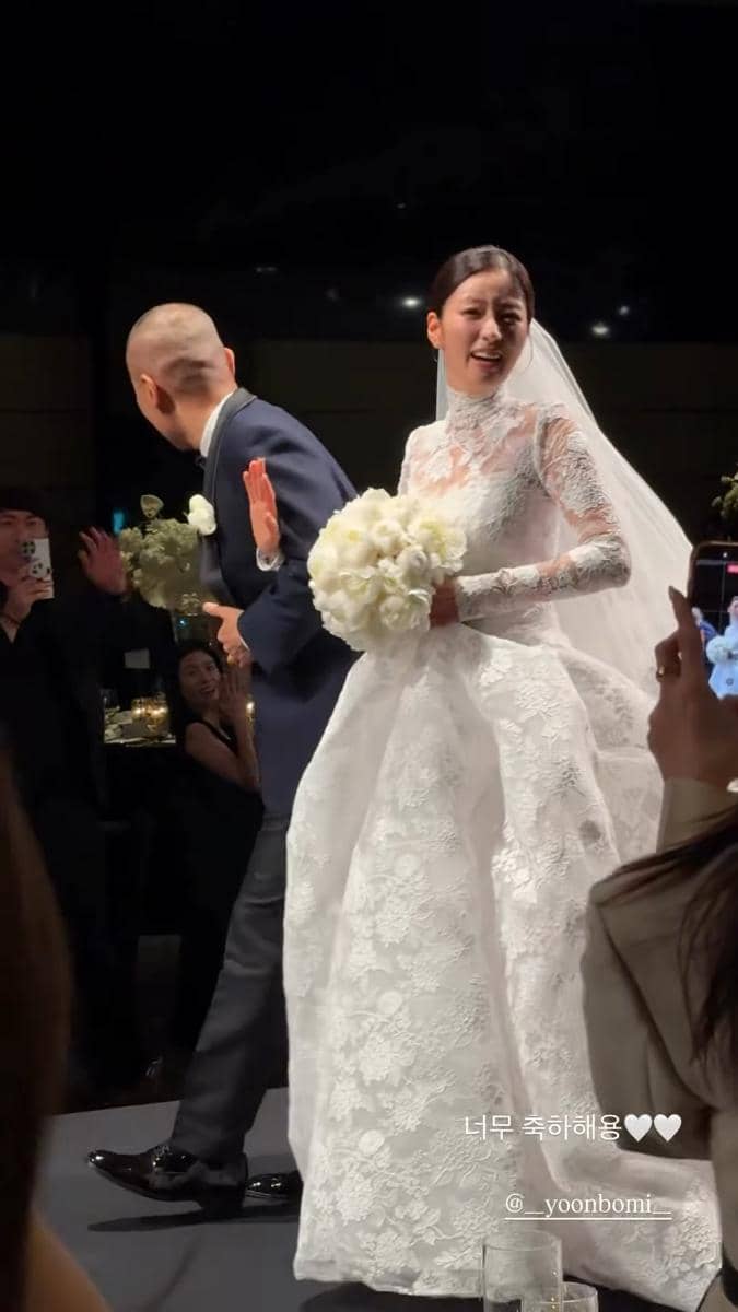 Seorang pengantin wanita mengenakan gaun putih renda panjang sambil memegang buket bunga, berdiri di samping mempelai pria di atas panggung pernikahan.
