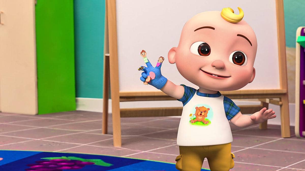 Karakter animasi bayi dari serial Cocomelon tersenyum sambil memegang boneka jari di ruang bermain berwarna cerah.