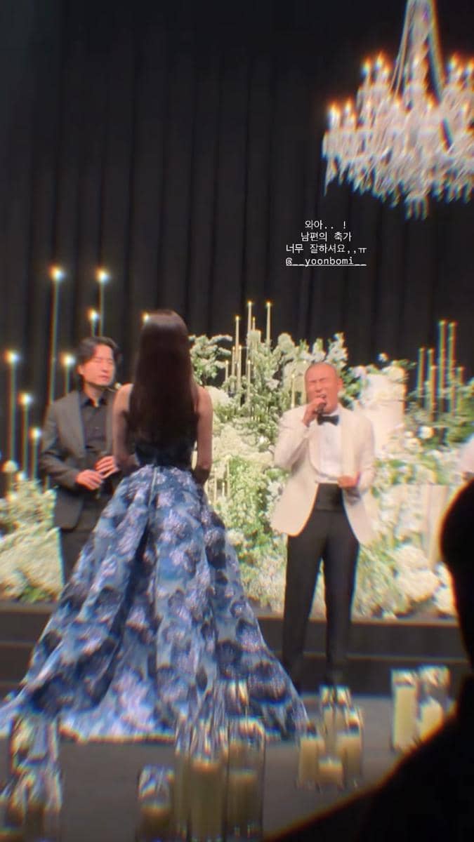 Seorang wanita mengenakan gaun biru bermotif bunga berdiri di depan dua pria di atas panggung dengan dekorasi bunga dan lilin di acara pernikahan.