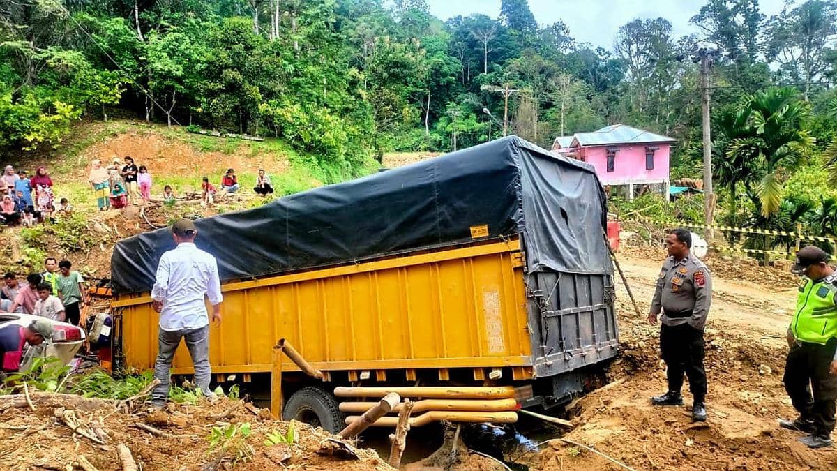 Truk berwarna kuning dengan terpal hitam tergelincir di tepi jalan tanah di Tapanuli Selatan, disaksikan warga dan petugas kepolisian.