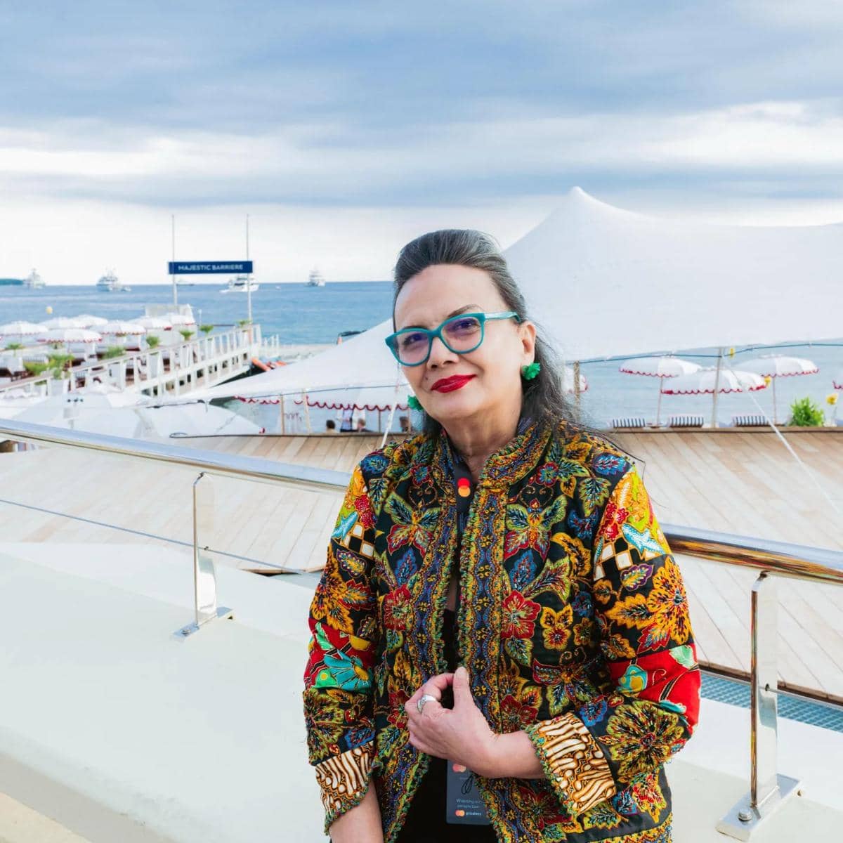 Seorang wanita mengenakan kacamata biru dan jaket bermotif batik warna-warni berpose di tepi pantai dengan latar laut dan tenda putih.