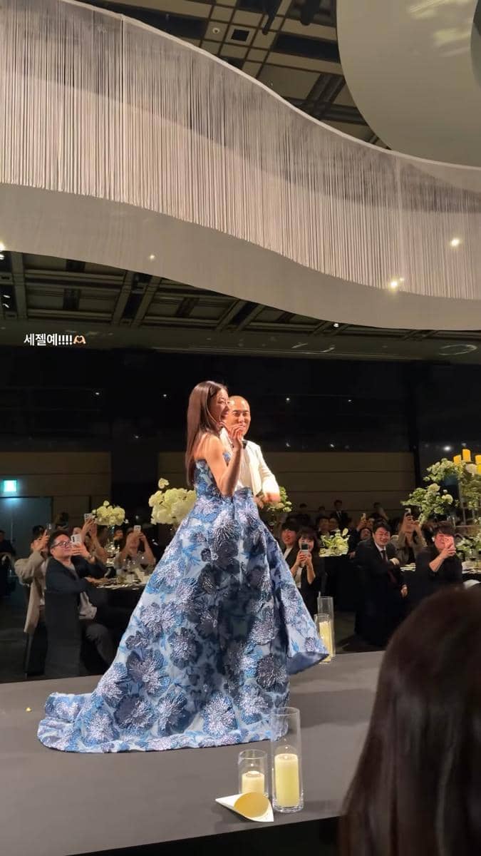 Seorang pengantin wanita mengenakan gaun biru bermotif bunga berjalan di panggung pernikahan bersama pasangannya di depan para tamu.