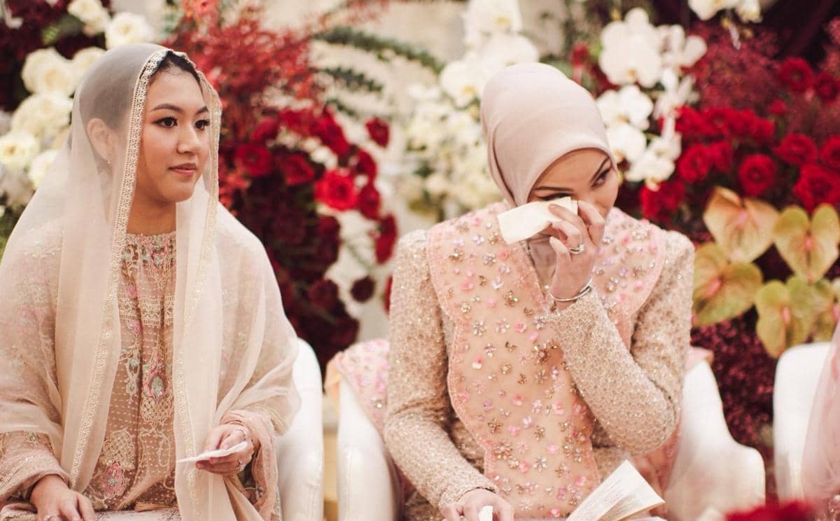 Dua perempuan mengenakan busana tradisional warna pastel duduk di acara pengajian dengan latar bunga merah dan putih.