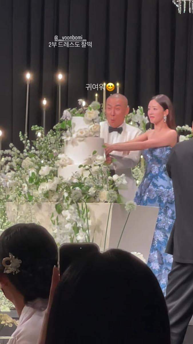 Pasangan pengantin memotong kue pernikahan bertingkat di atas meja yang dihiasi bunga putih dengan latar tirai hitam elegan.