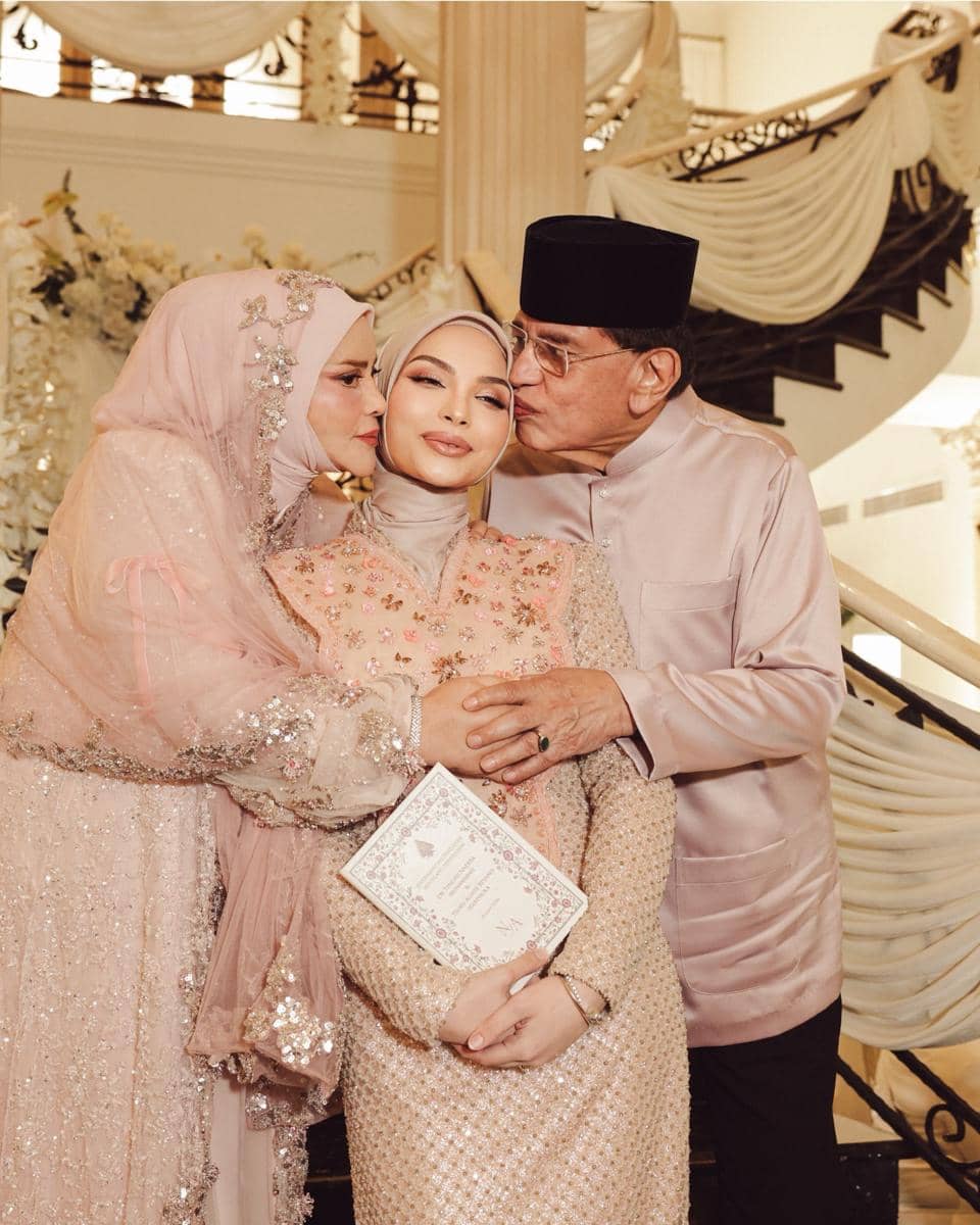 Seorang wanita berhijab berbusana tradisional warna pastel berpose bersama dua orang lanjut usia yang menciumnya di pipi di acara pengajian jelang pernikahan.