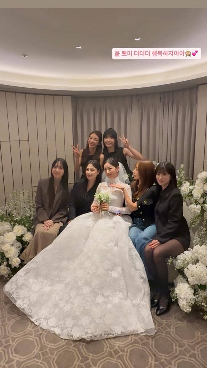 Seorang pengantin wanita mengenakan gaun putih duduk di tengah bersama teman-temannya yang berpose ceria di ruangan berhias bunga putih.