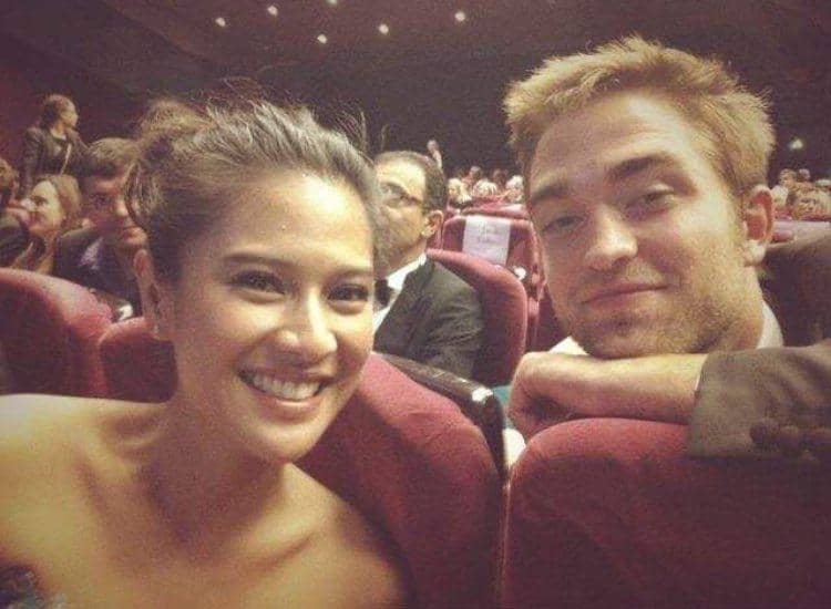 Seorang perempuan tersenyum bersama seorang pria di kursi teater berwarna merah dengan suasana ramai di latar belakang.