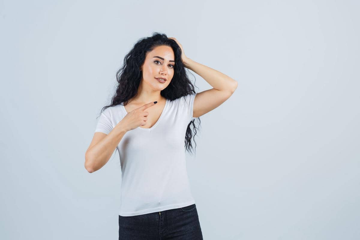 Perempuan berambut panjang mengenakan kaus putih menyentuh rambutnya sambil menunjuk ke arah kepala dengan latar abu-abu polos.