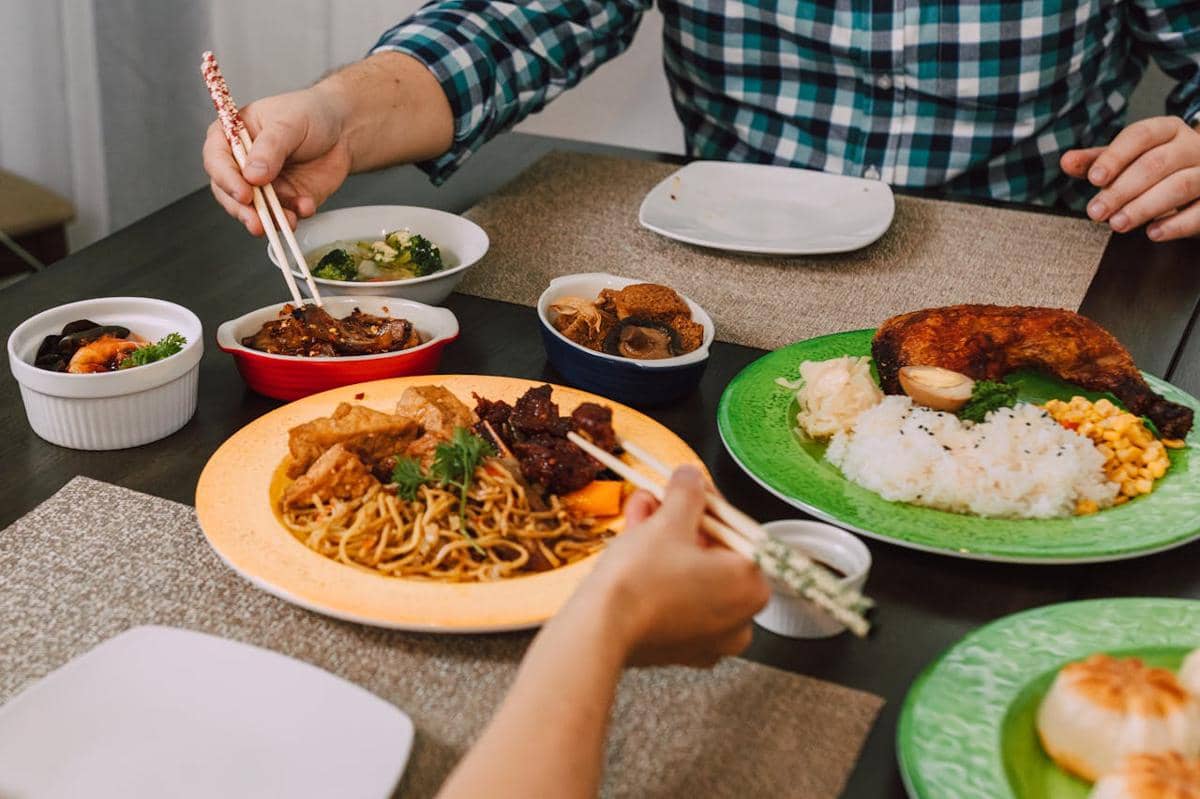Dua orang sedang menikmati hidangan Asia di meja makan dengan berbagai menu seperti nasi, ayam panggang, mie, dan sayuran.