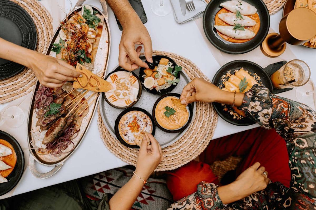 Beberapa orang menikmati hidangan bersama di meja makan dengan berbagai makanan seperti saus, sate, dan roti pita di atas piring hitam.
