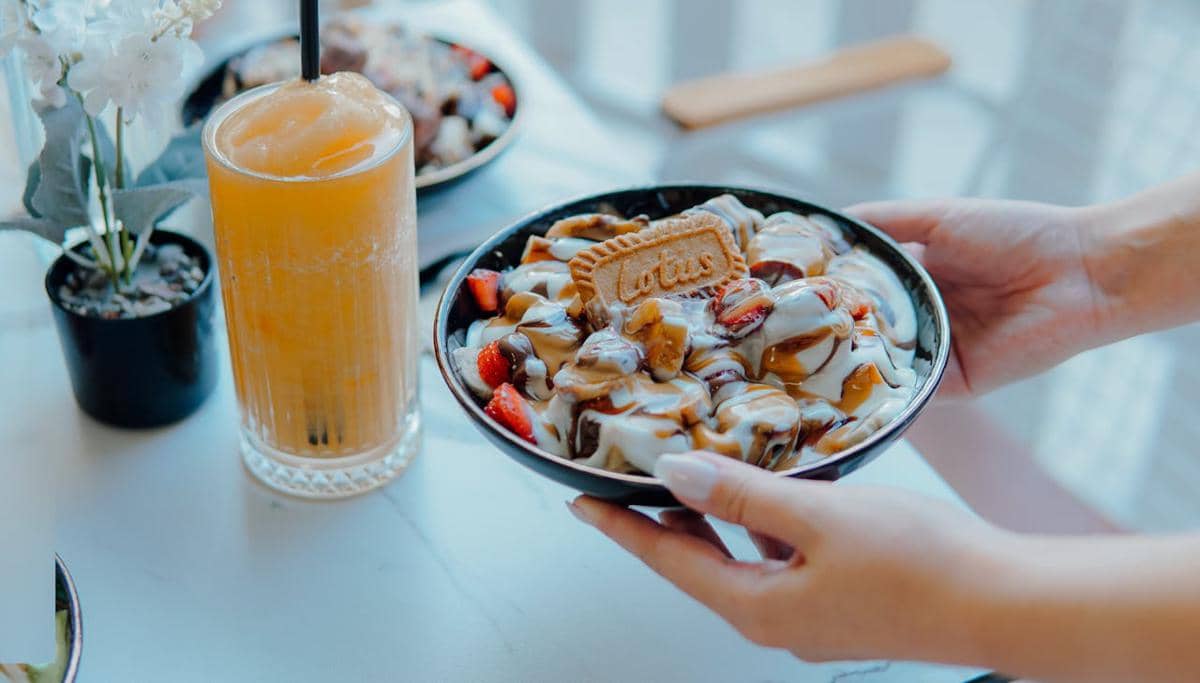 Semangkuk makanan manis berisi potongan buah dan biskuit dengan saus krim dan karamel di atas meja bersama segelas minuman dingin.