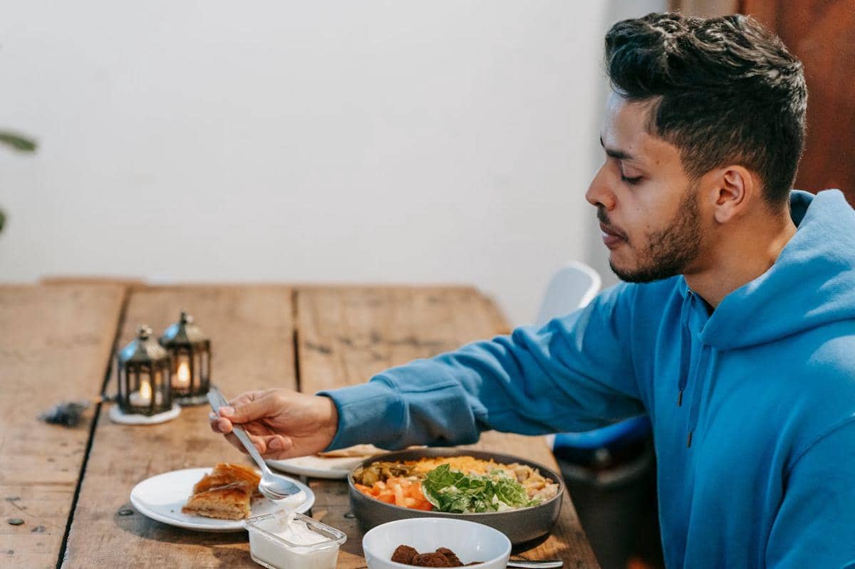 Seorang pria mengenakan hoodie biru sedang makan di meja kayu dengan berbagai hidangan dan lentera kecil di atas meja.