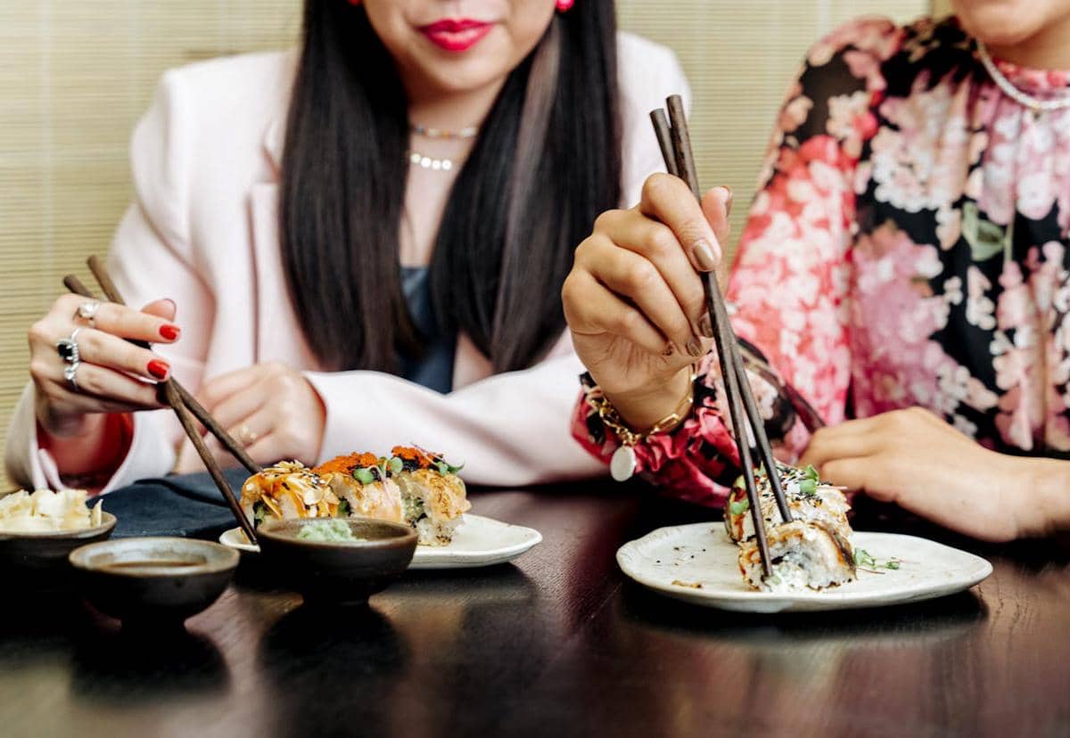 Dua wanita mengenakan pakaian berwarna cerah menikmati hidangan sushi dengan sumpit di atas meja restoran.