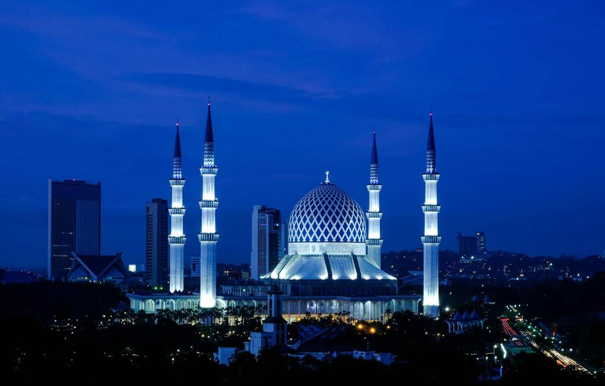 Masjid megah di Shah Alam dengan kubah biru besar dan empat menara tinggi yang diterangi cahaya malam di bawah langit biru gelap.