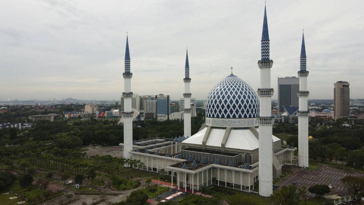 Masjid besar dengan kubah biru dan empat menara tinggi berdiri megah di tengah kota Shah Alam dengan latar gedung modern.