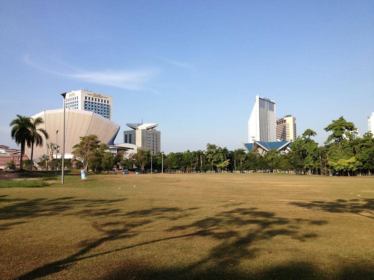 Pemandangan kota Shah Alam dengan gedung-gedung modern di latar belakang dan area lapangan rumput luas di bawah langit cerah.