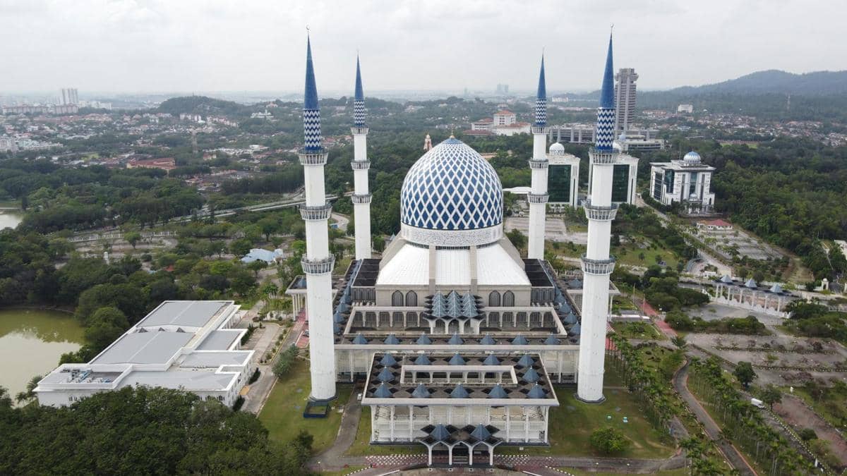 Masjid megah di Shah Alam dengan kubah biru besar dan empat menara tinggi dikelilingi pepohonan serta bangunan kota.