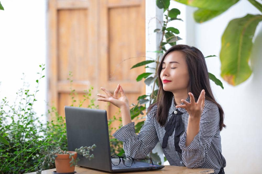 Perempuan duduk di depan laptop dengan mata terpejam dan tangan dalam posisi meditasi sambil mengatur napas di ruang terbuka hijau.