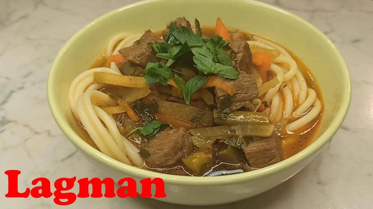 Semangkuk lagman berisi mi tebal dengan potongan daging sapi, sayuran, dan kuah berwarna merah kecokelatan, dihiasi daun hijau segar.