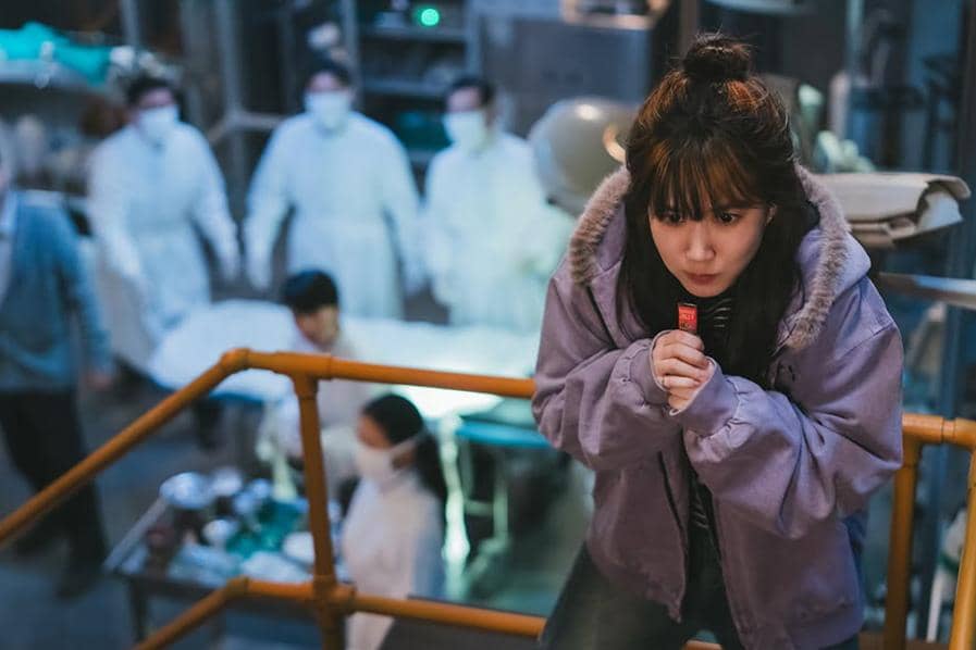 Seorang wanita mengenakan jaket ungu berdiri di atas tangga logam sambil memegang benda kecil, dengan beberapa orang berseragam medis di latar belakang.