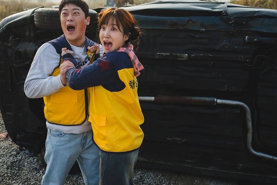 Dua orang mengenakan rompi kuning tampak terkejut di depan mobil yang terbalik di lokasi luar ruangan pada adegan drama The WONDERfools.