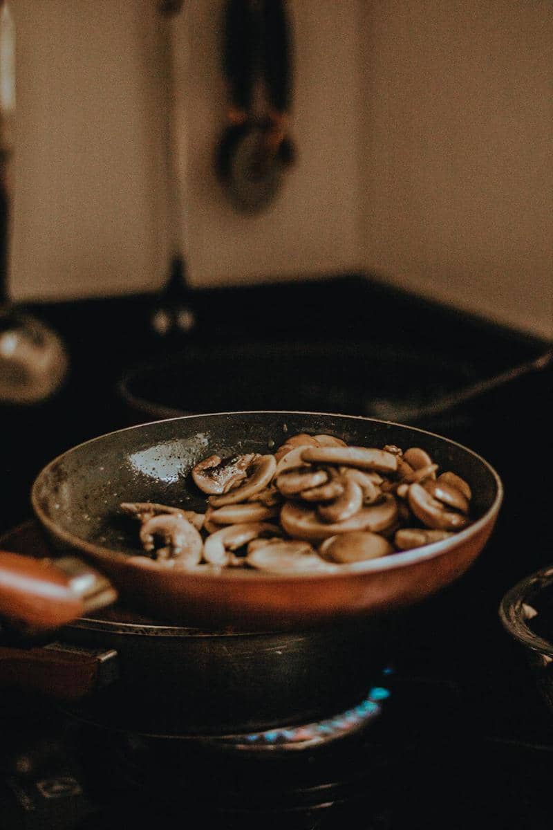 Irisan jamur sedang ditumis di atas wajan di dapur dengan api kompor menyala, menciptakan suasana hangat saat memasak.