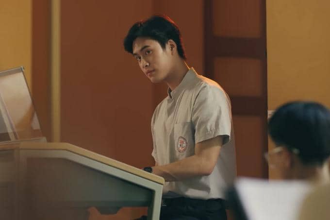 Seorang siswa berseragam abu-abu duduk di depan keyboard di ruang kelas dengan pencahayaan hangat, menatap ke arah seseorang.