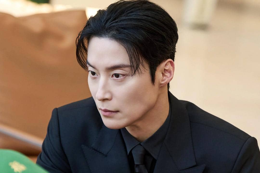 Seorang pria berambut hitam mengenakan setelan jas hitam tampak serius dalam adegan drama My Royal Nemesis.