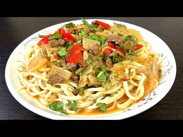 Sepiring lagman dengan mi tebal disajikan bersama potongan daging sapi, paprika merah, sayuran, dan taburan daun ketumbar segar.