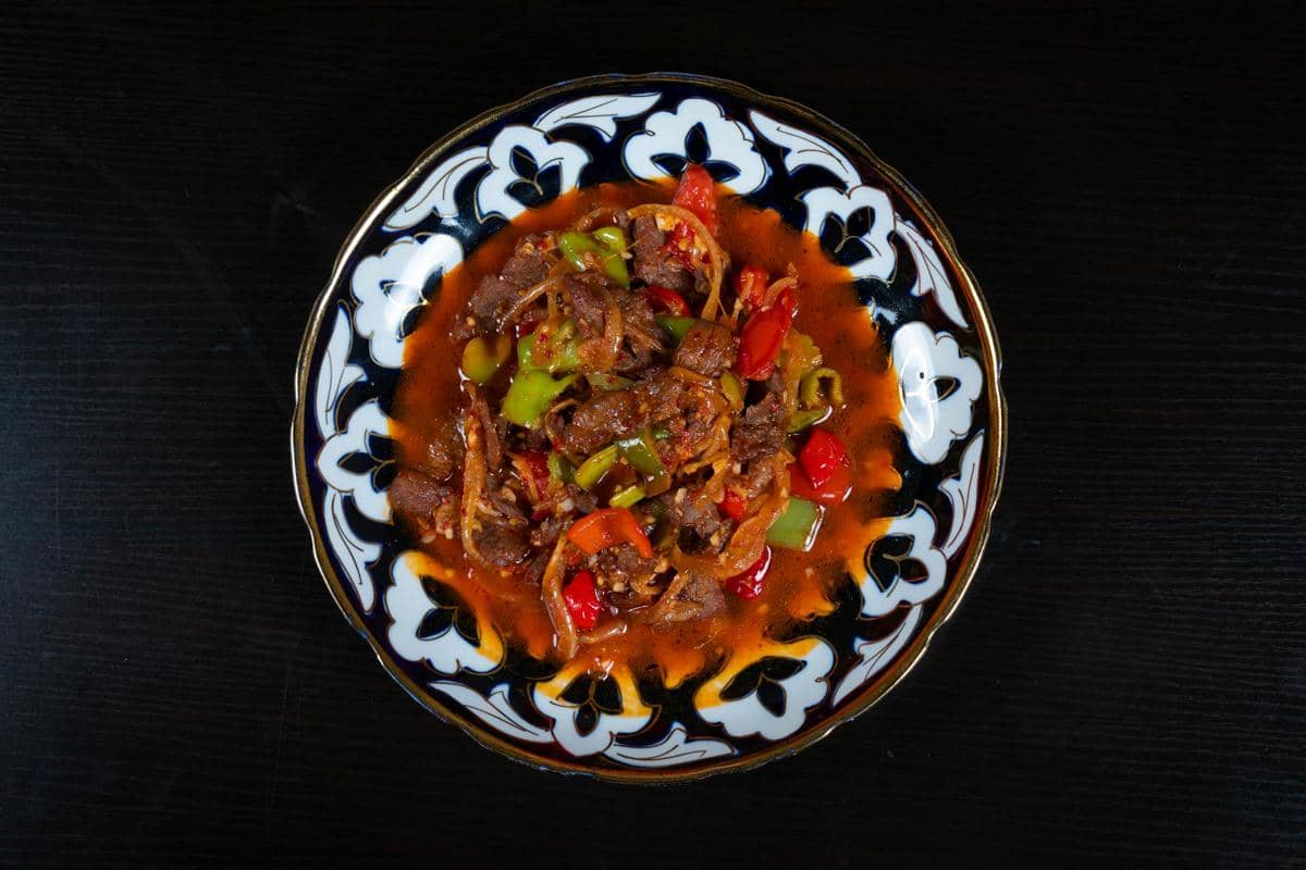 Semangkuk lagman dengan potongan daging sapi, paprika merah dan hijau, serta saus merah kental disajikan di atas piring bermotif tradisional.