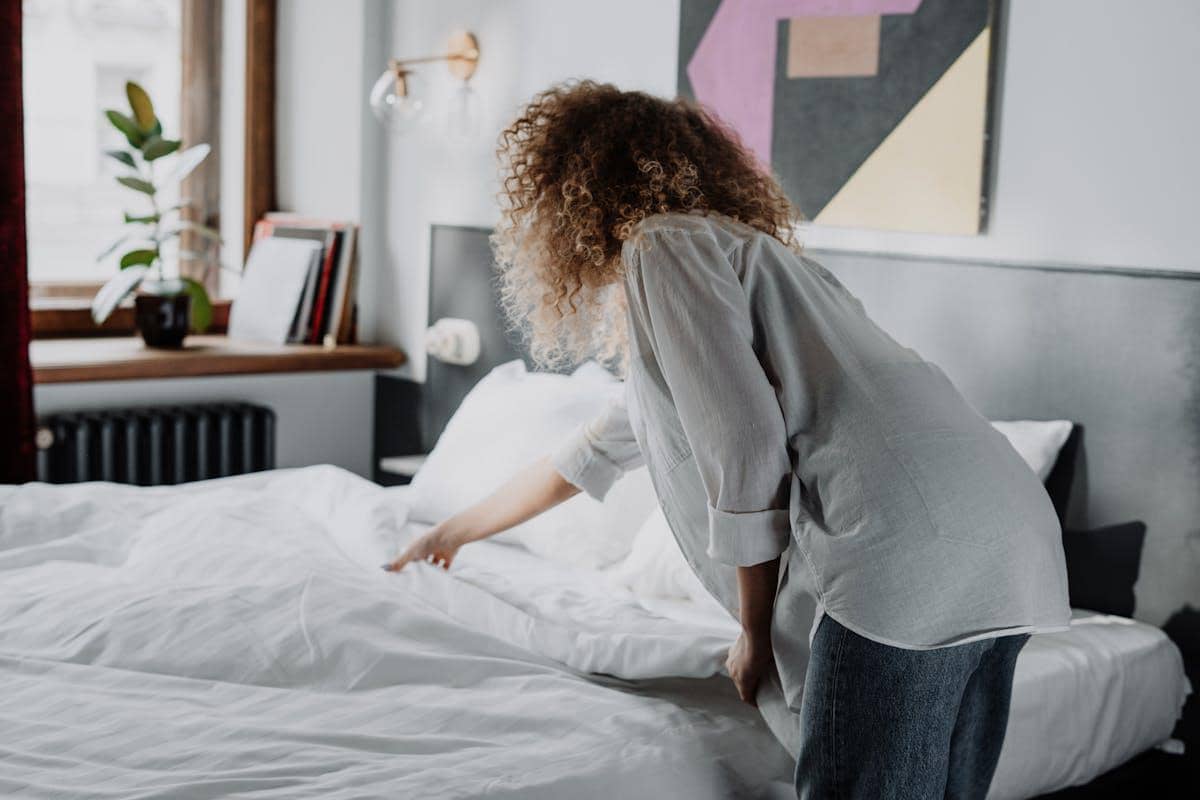 Seorang perempuan sedang merapikan tempat tidur dengan seprai putih di kamar yang terang dan rapi dengan dekorasi minimalis.