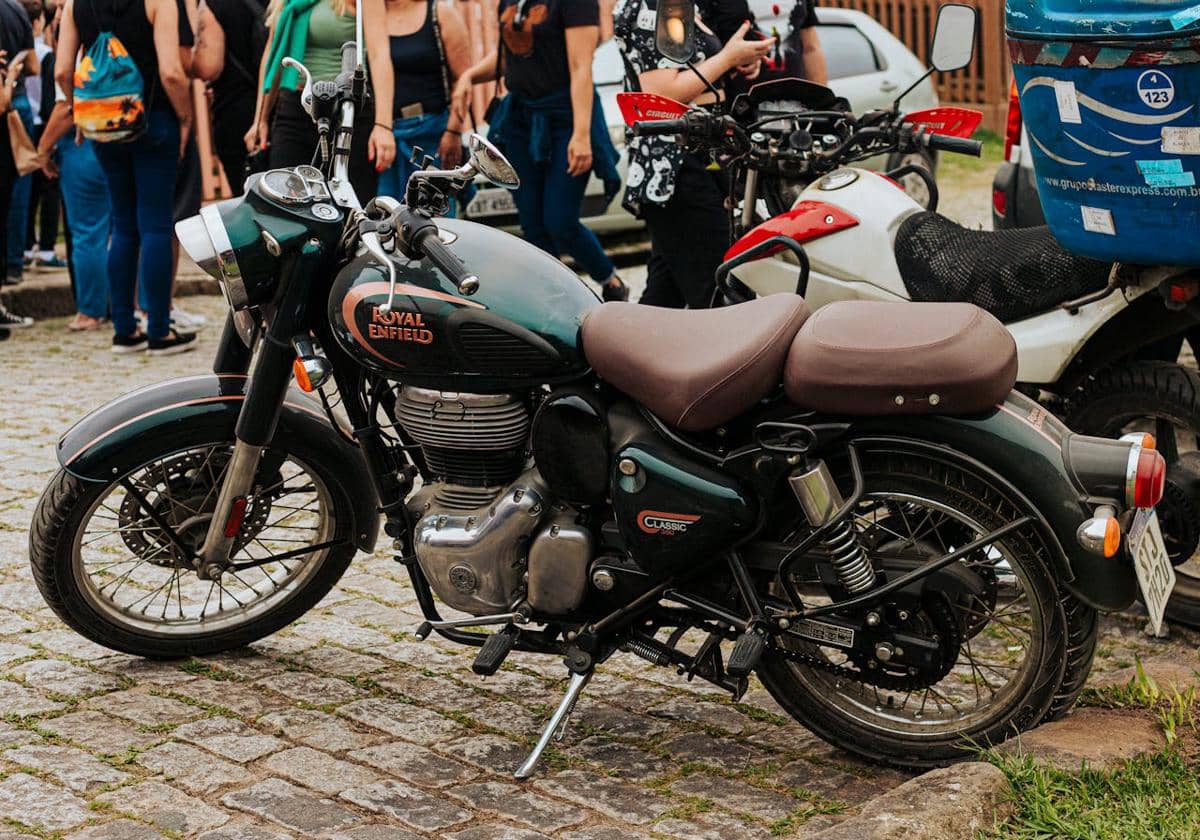 Motor bergaya retro modern berwarna hijau tua dengan jok cokelat terparkir di jalan berbatu, dikelilingi beberapa orang di latar belakang.