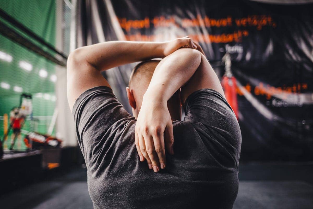 Seseorang melakukan peregangan lengan di dalam ruangan olahraga dengan latar belakang peralatan fitness dan dinding bertulisan.