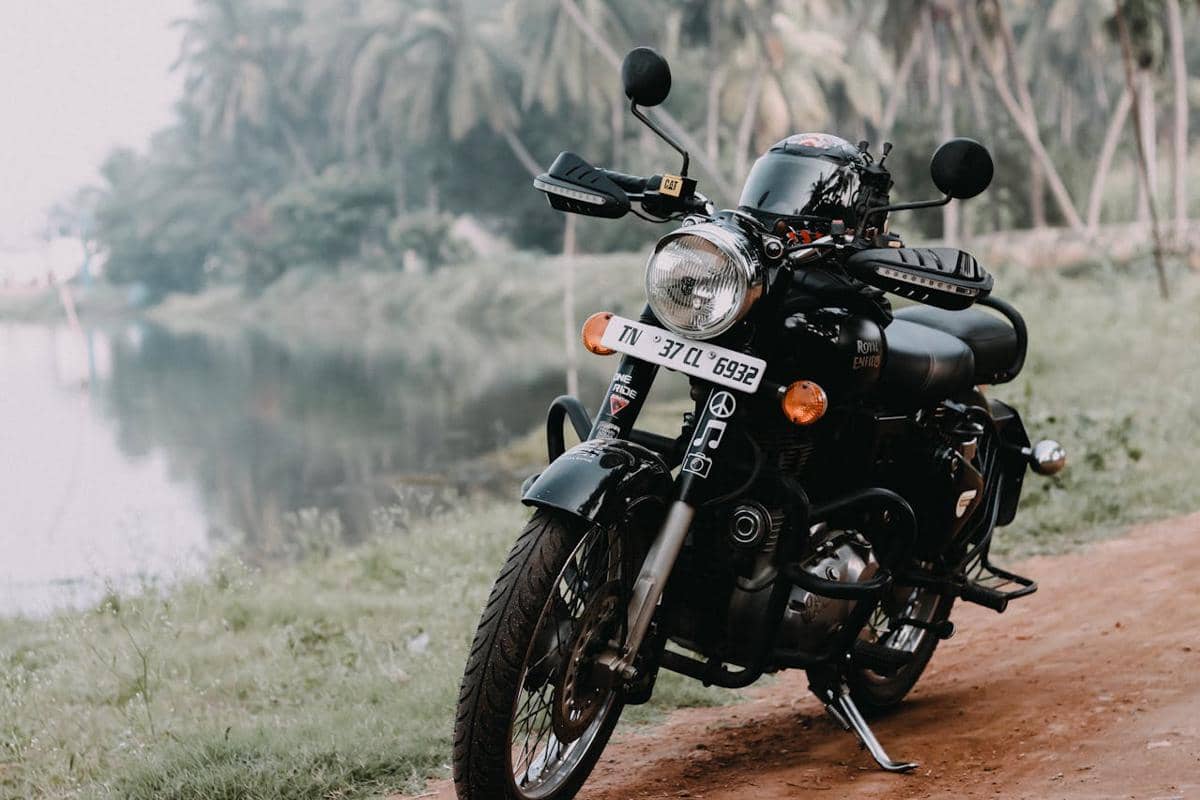 Motor retro modern berwarna hitam terparkir di jalan tanah dekat sungai dengan latar belakang pepohonan hijau.