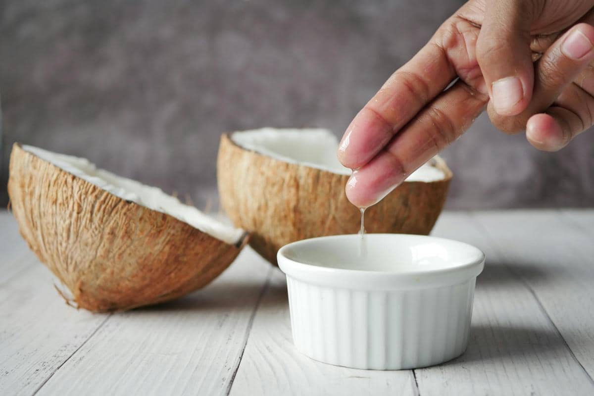 Tangan mengambil minyak kelapa dari wadah kecil putih dengan dua belahan kelapa di atas meja kayu berwarna terang.