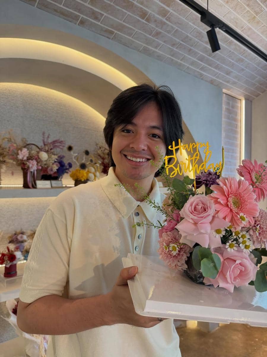 Seorang pria tersenyum sambil memegang rangkaian bunga berwarna pink dengan hiasan tulisan Happy Birthday di ruangan bernuansa hangat.