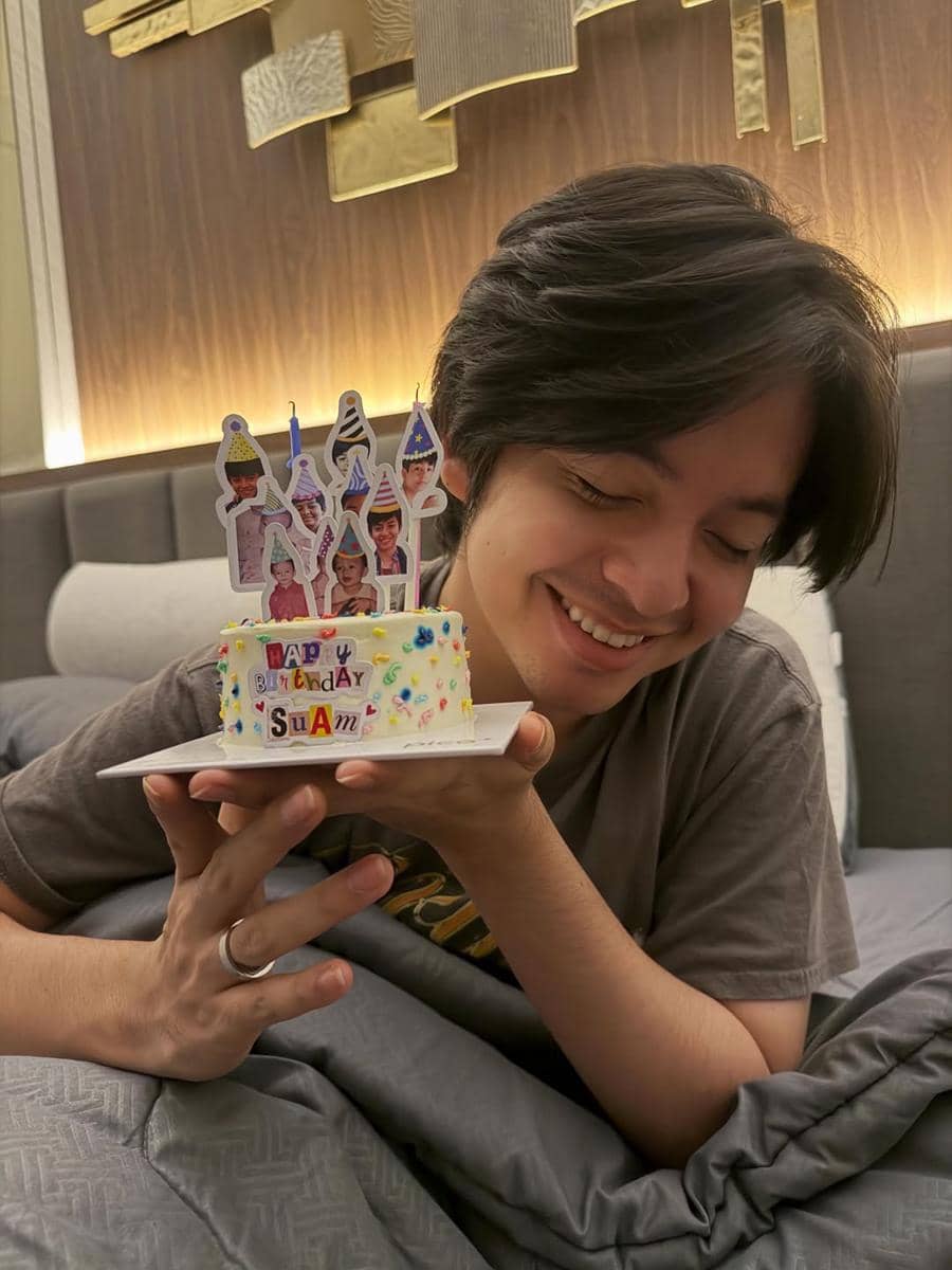 Seorang pria tersenyum sambil memegang kue ulang tahun kecil berdekorasi warna-warni di atas tempat tidur dengan latar kamar modern.